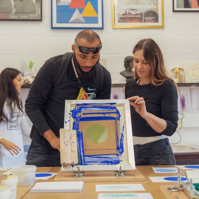 Two people screen printing together, pulling a blue and gold print.