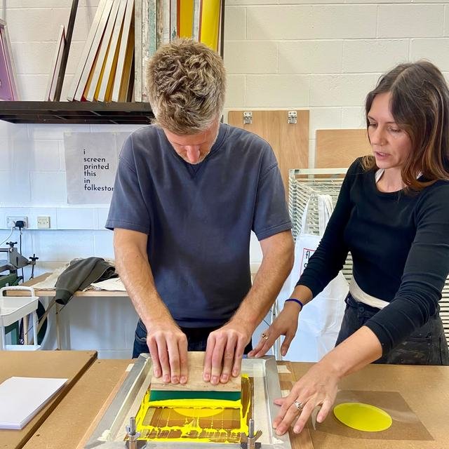 Two people pulling a yellow and green print together at the studio bench.