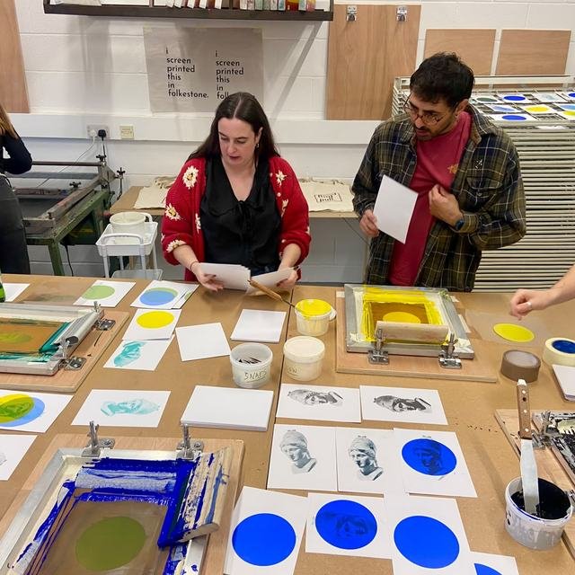 A group of four at the print table, surrounded by coloured ink and finished prints.